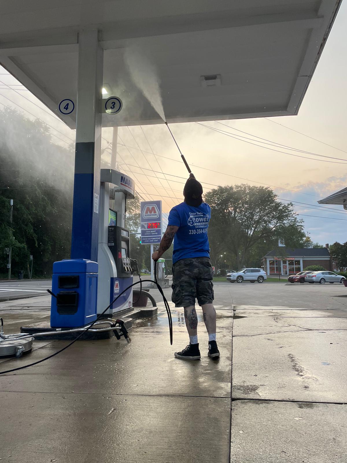 A person in blue overalls and yellow boots uses a pressure washer to clean a garage door, with a water hose connected to the machine. The setting features a modern building, and there are potted plants nearby.